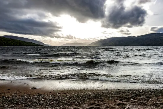A wide landscape photo shows dark, choppy water with white-capped waves breaking on a rocky and sandy shore. Distant hills…