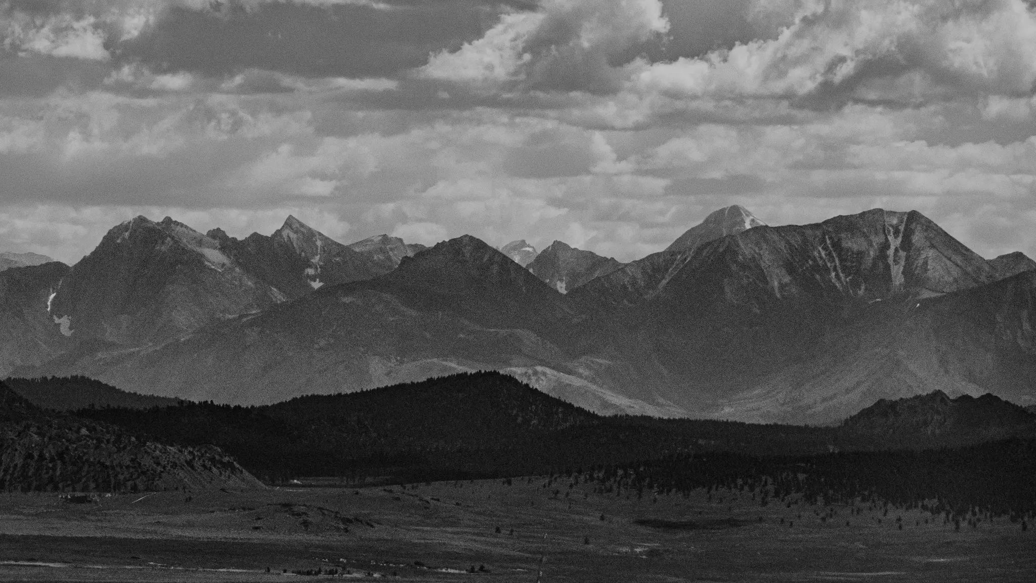 Une photo de paysage panoramique en noir et blanc d'une chaîne de montagnes sous un ciel nuageux, avec des collines boisées