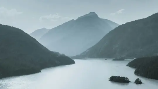 A wide landscape photo of a calm, blue-grey lake surrounded by steep, forested mountains under a hazy sky. Small islands…