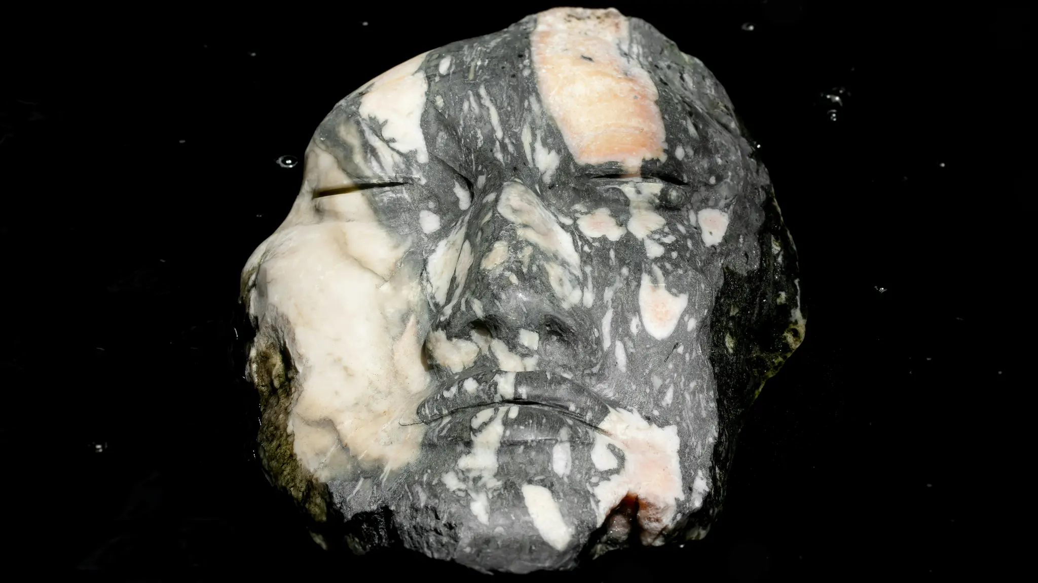 Gros plan d'une tête de pierre sculptée aux yeux fermés, avec un motif moucheté de blanc, de gris et de rose pâle/orange.