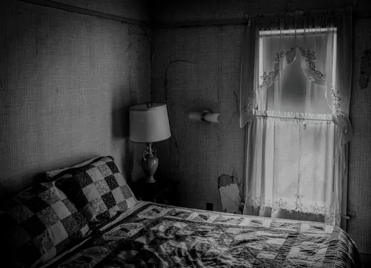 A black and white photo of a bedroom with a quilted bed, a lamp on a nightstand, and a window with sheer curtains.