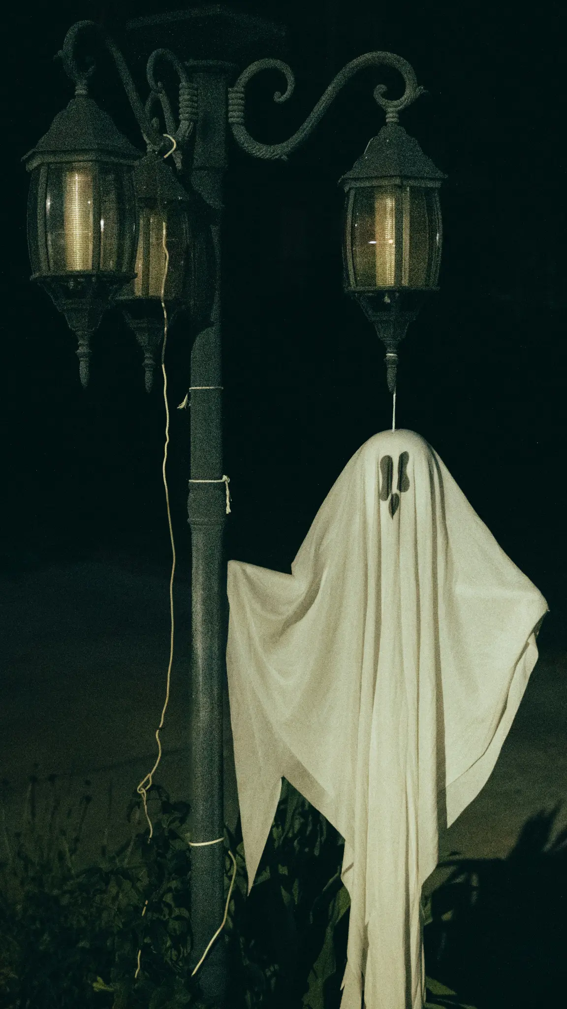Une décoration fantôme blanche en forme de drap avec des yeux et une bouche noirs est suspendue à un lampadaire sombre avec