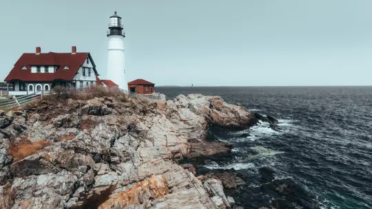 A white lighthouse and a white building with a red roof stand on a rugged, rocky coastline. The dark blue ocean stretches…