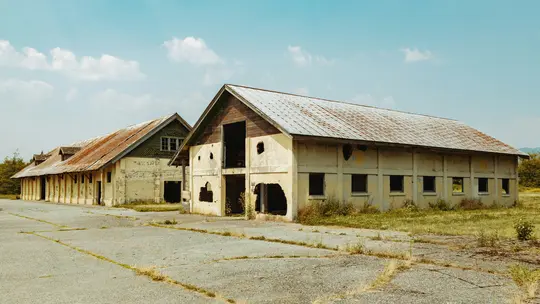 Two long, abandoned buildings with yellowing, crumbling walls and heavily rusted metal roofs stand under a partly cloudy sky.