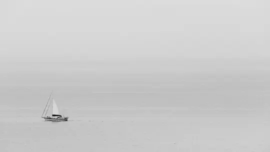 Čiernobiela širokouhlá fotografia plachetnice s bielou plachtou na pokojnej vode pod hmlistou oblohou.