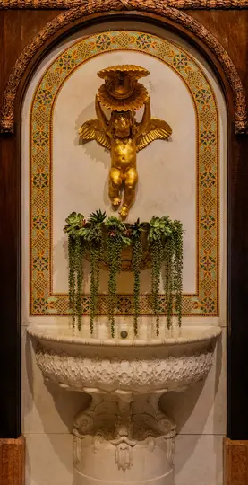 Detailná nástenná fontána so zlatým anjelikom držiacim mušľu, nad kvetináčom so sukulentmi a vyrezávanou mramorovou nádobou.