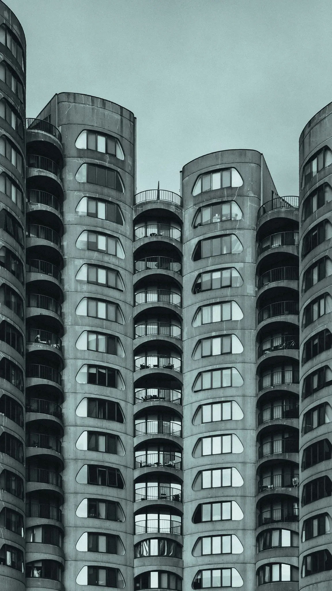 Una foto monocromatica, scattata dal basso, di diversi edifici brutalisti in cemento. Presentano balconi arrotondati e…