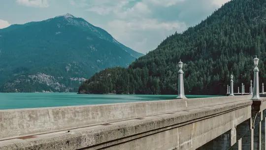 A concrete dam or bridge with decorative lampposts extends across the foreground, overlooking a vibrant turquoise lake.