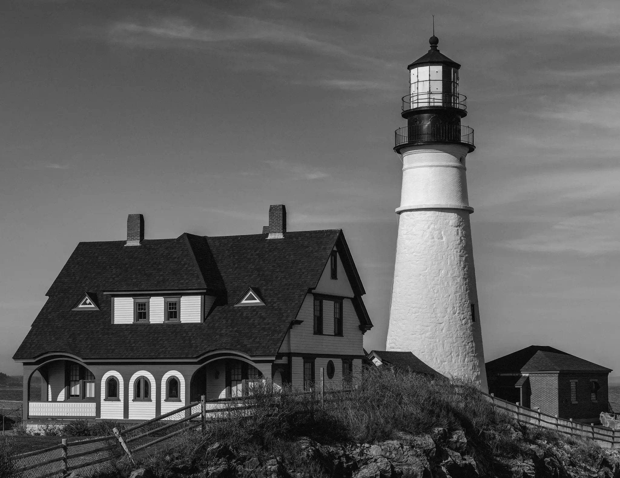 Une photo en noir et blanc montre un phare blanc et haut à côté d'une grande maison au toit sombre, tous deux sur une colline