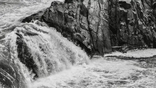 A black and white photo shows a powerful waterfall crashing over dark, textured rocks into a frothy river below.