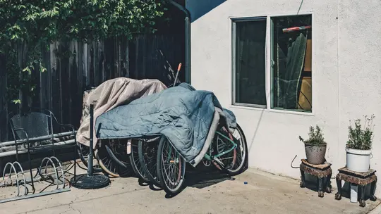 Several bicycles are covered with a blue blanket and a tan tarp, parked against a wooden fence and a white wall.