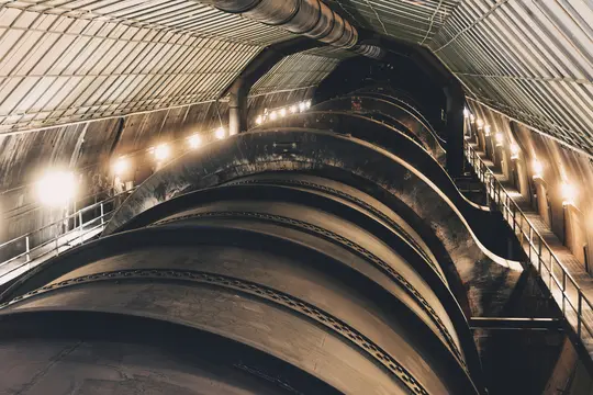 A low-angle shot inside a large industrial building, showing a massive, dark, curved pipe-like structure in the foreground.