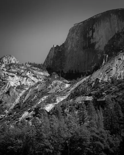 A black and white photo showing a massive, textured rock face dominating the upper right, with a dense forest at its base.
