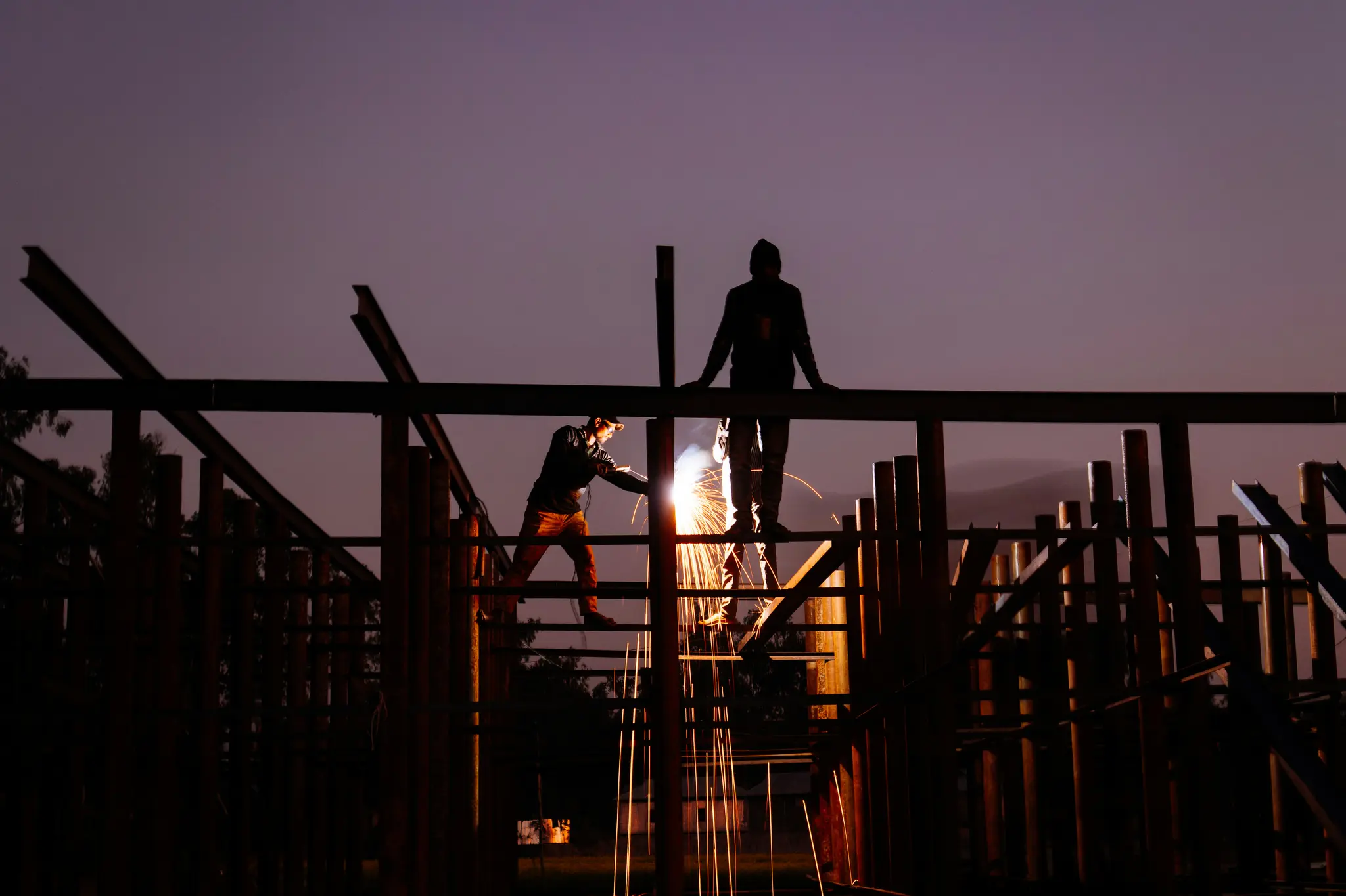 Deux ouvriers du bâtiment en silhouette sur un ciel crépusculaire violet-gris. Un ouvrier soude des poutres métalliques