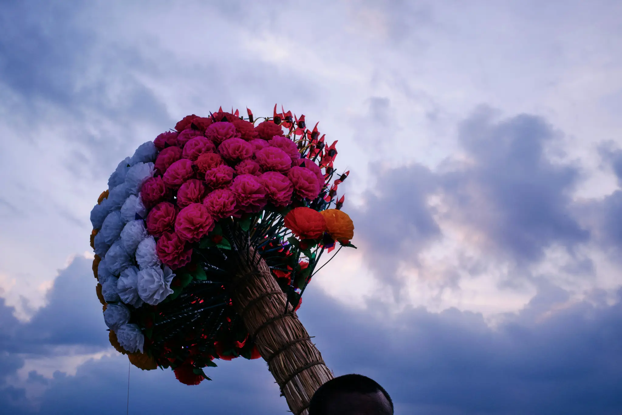 En stor, färgstark bukett av konstgjorda blommor, främst rosa och vita med lite orange, hållen mot en molnig himmel