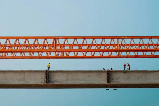 Fem byggnadsarbetare på en betongbalk under ett stort orange metallfackverk mot en klarblå himmel. Arbetarna är…