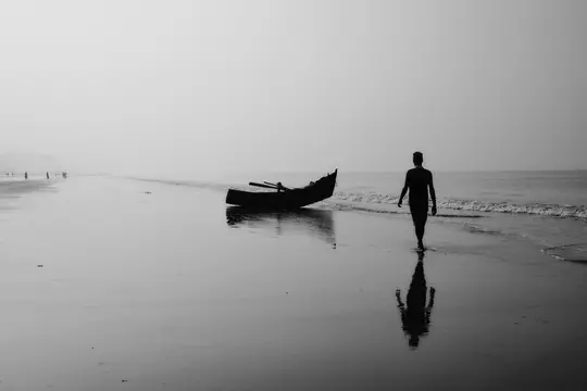 Černobílá fotografie muže v siluetě kráčejícího po pláži vedle malé lodi, s mlhavou oblohou a oceánem.