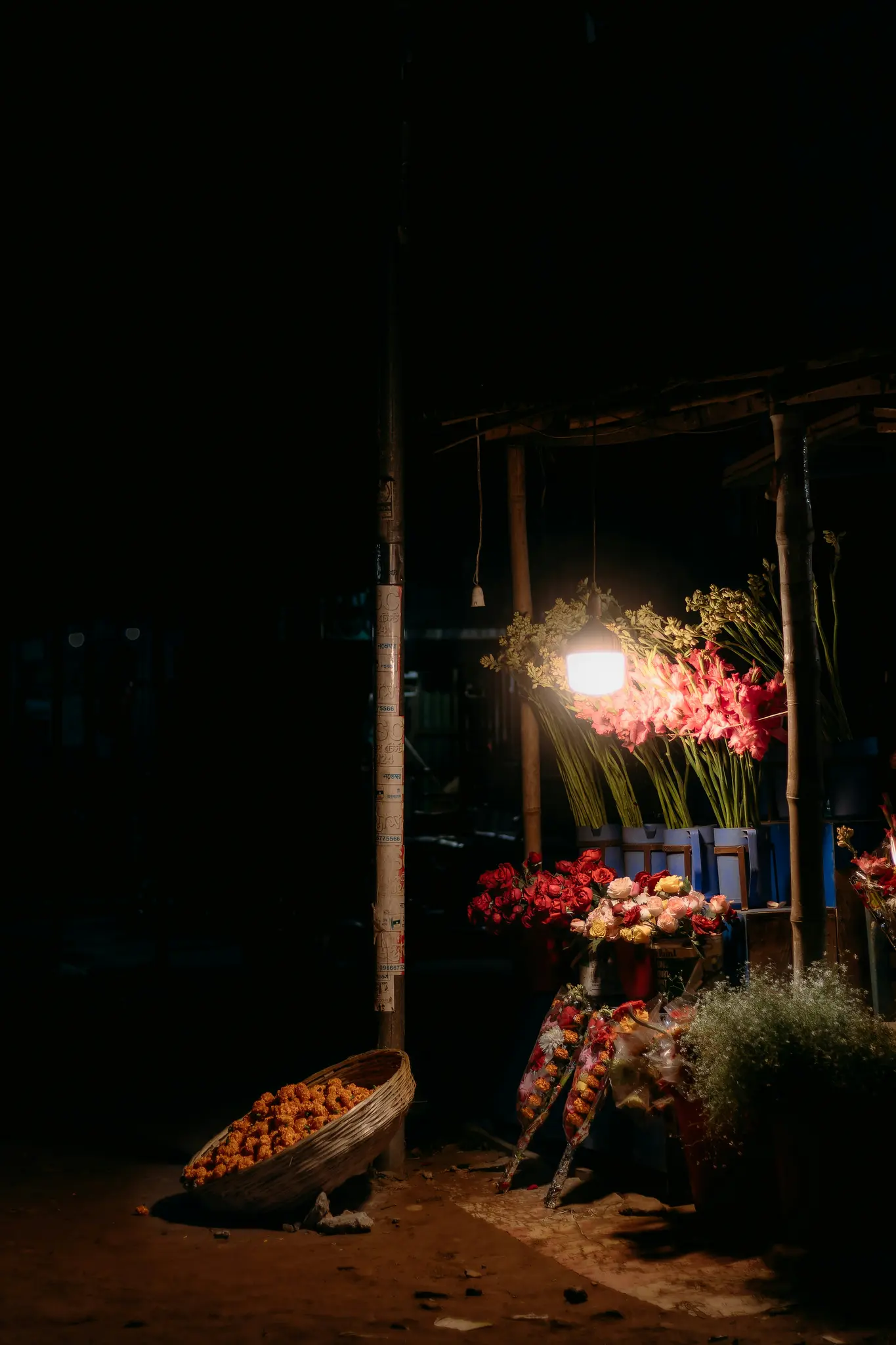 En svagt upplyst nattscéna visar ett blomsterstånd med olika buketter under en enda glödande glödlampa. En korg med orange…