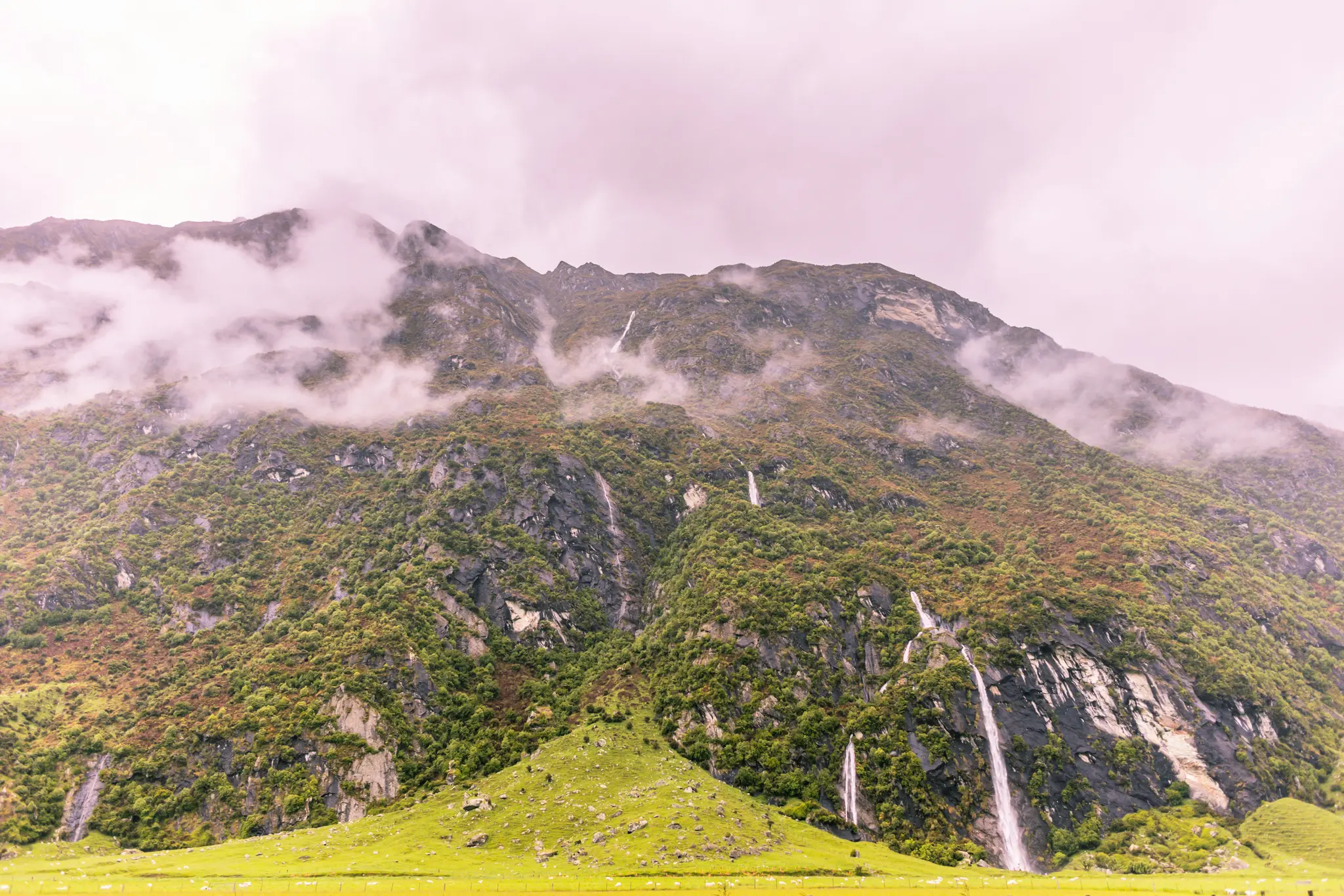 A vast mountain covered in green foliage and grey rock, with several thin waterfalls flowing down. Clouds cling to the…