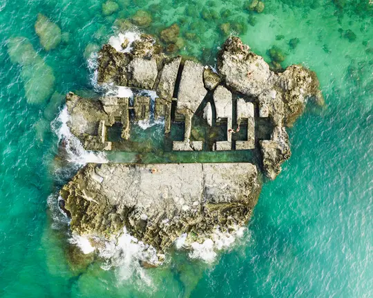 Una vista aerea, dall'alto, di un'isola rocciosa con strutture geometriche in cemento, circondata da un limpido oceano