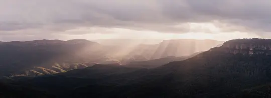 Ett brett landskapsfoto visar solstrålar som bryter igenom mörka moln och lyser upp delar av en vidsträckt, skogbevuxen
