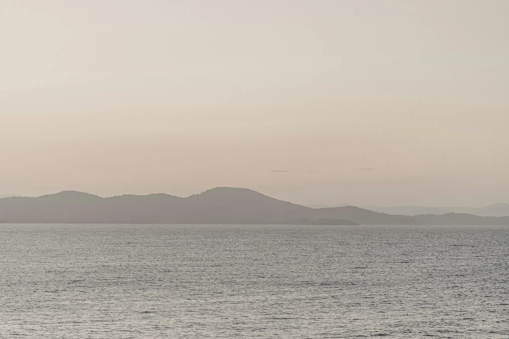 Una foto di paesaggio minimalista che mostra acqua calma e grigia in primo piano e strati di colline scure e nebbiose sotto