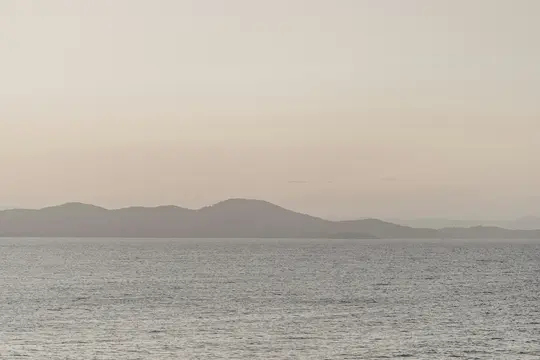 Ein minimalistisches Landschaftsfoto, das ruhiges, graues Wasser im Vordergrund und Schichten von dunstigen, dunklen Hügeln