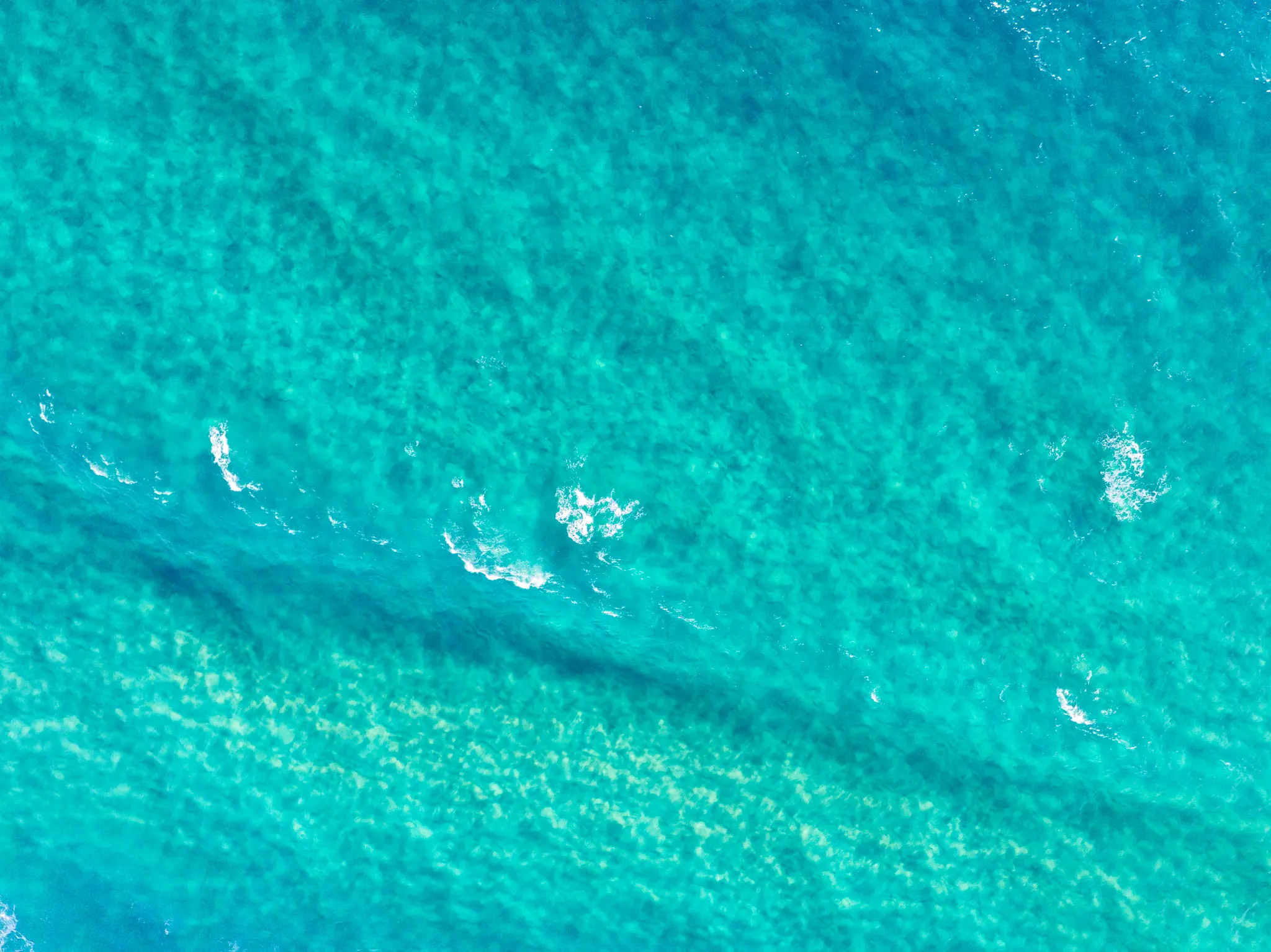 An aerial, top-down view of clear turquoise ocean water with small white waves and visible light sand patterns beneath.