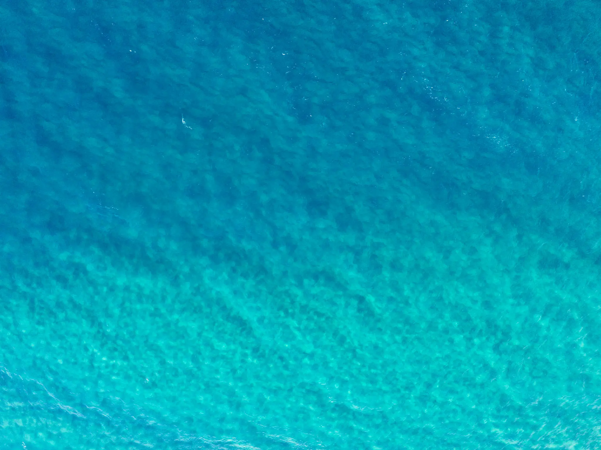An aerial photograph shows clear ocean water, transitioning from deep blue at the top to lighter turquoise at the bottom.