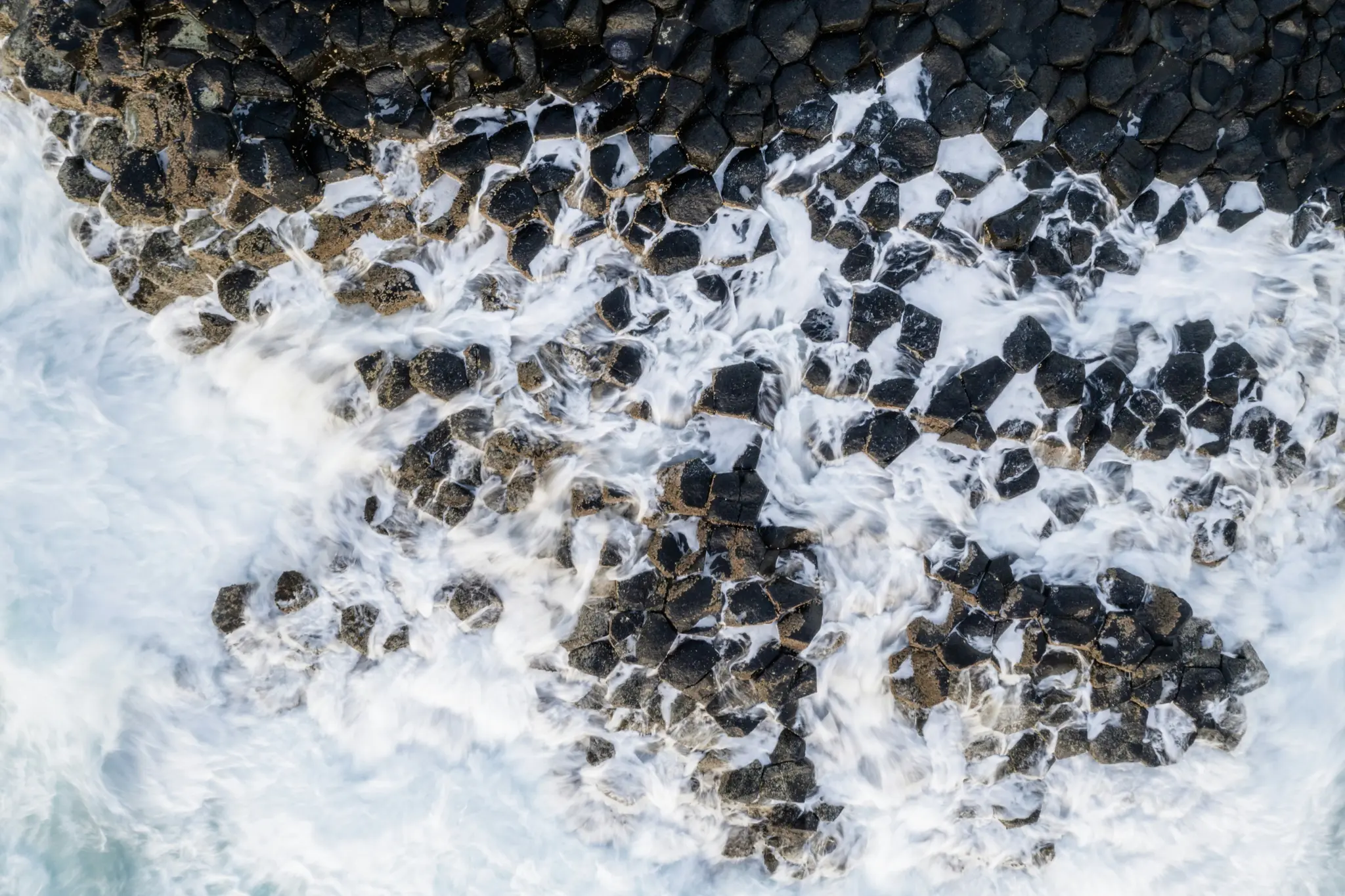 An aerial view captures white ocean waves crashing over dark, hexagonal basalt columns, creating a dynamic contrast.