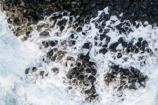 An aerial view captures white ocean waves crashing over dark, hexagonal basalt columns, creating a dynamic contrast.