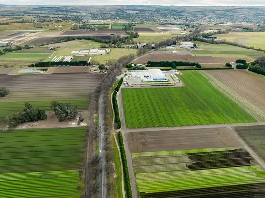 Una foto aerea di un paesaggio rurale con una lunga strada, campi agricoli verdi e marroni e un grande edificio industriale