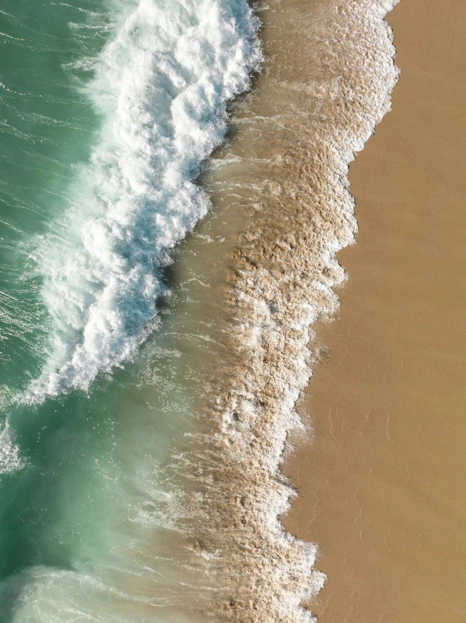 Widok z lotu ptaka, z góry, pokazuje turkusową falę oceaniczną łamiącą się na beżowej piaszczystej plaży, tworząc białą