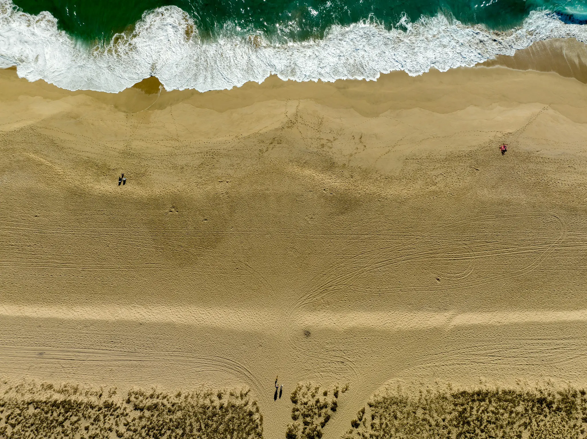 Et luftfoto, set ovenfra, af en bred sandstrand. Grønt havvand med hvide, skummende bølger slår ind mod det lysebrune sand.