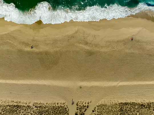 Eine Luftaufnahme, von oben, eines breiten Sandstrandes. Grünes Meerwasser mit weißen, schaumigen Wellen bricht sich auf dem