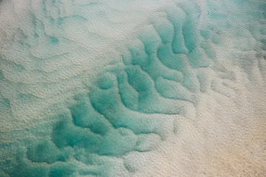 An aerial shot shows clear, shallow turquoise water revealing intricate ripple patterns on a light sandy seabed.