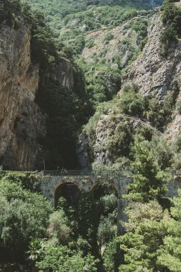 Eine Steinbrücke mit mehreren Bögen überspannt eine tiefe, baumgefüllte Schlucht zwischen steilen, felsigen Berghängen unter