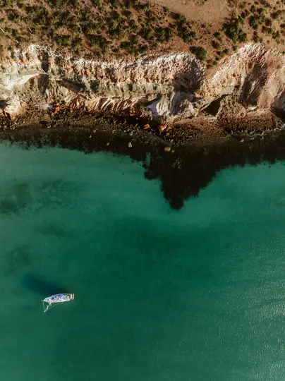 Una ripresa aerea cattura una barca a vela bianca in vibranti acque turchesi, ancorata vicino a una costa aspra con scogliere