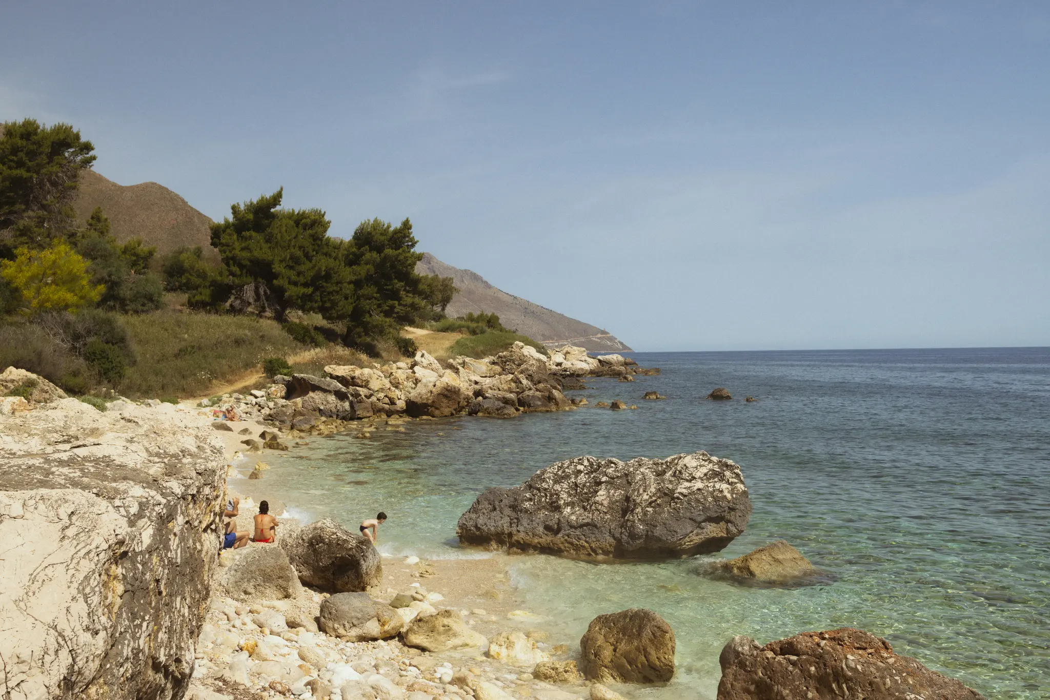 A wide landscape photo of a rocky beach with clear turquoise water. A few people are on the beach, backed by a green…