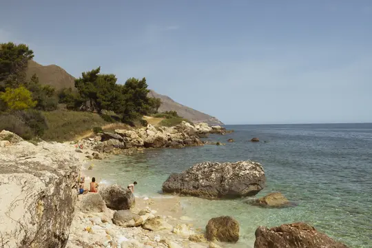 A wide landscape photo of a rocky beach with clear turquoise water. A few people are on the beach, backed by a green…