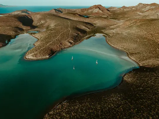 An aerial view shows a tranquil bay with three small white sailboats on vibrant turquoise water, surrounded by dry, brown…
