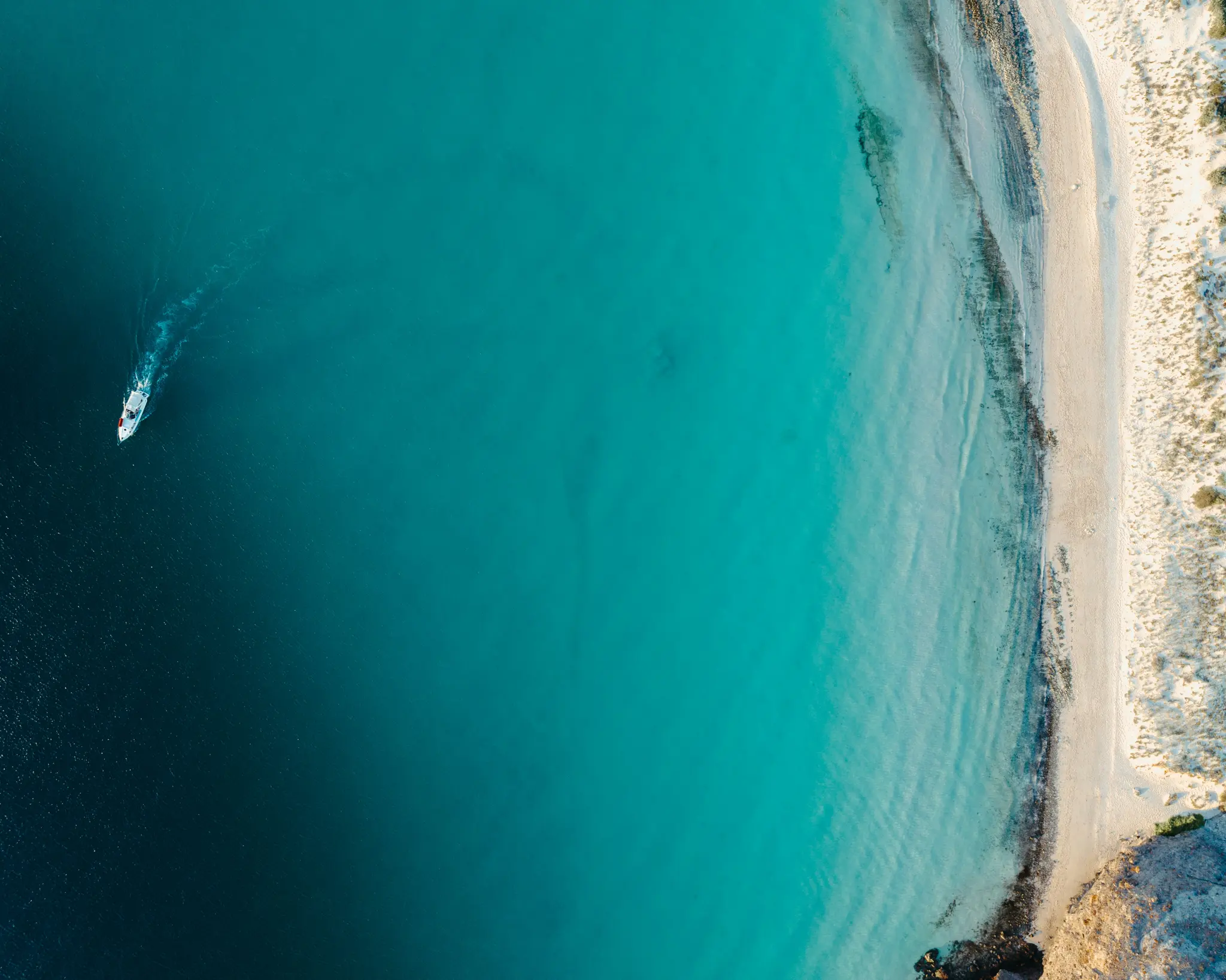 An aerial shot shows a small white boat with a red stripe moving across deep turquoise water, leaving a wake. To the right…