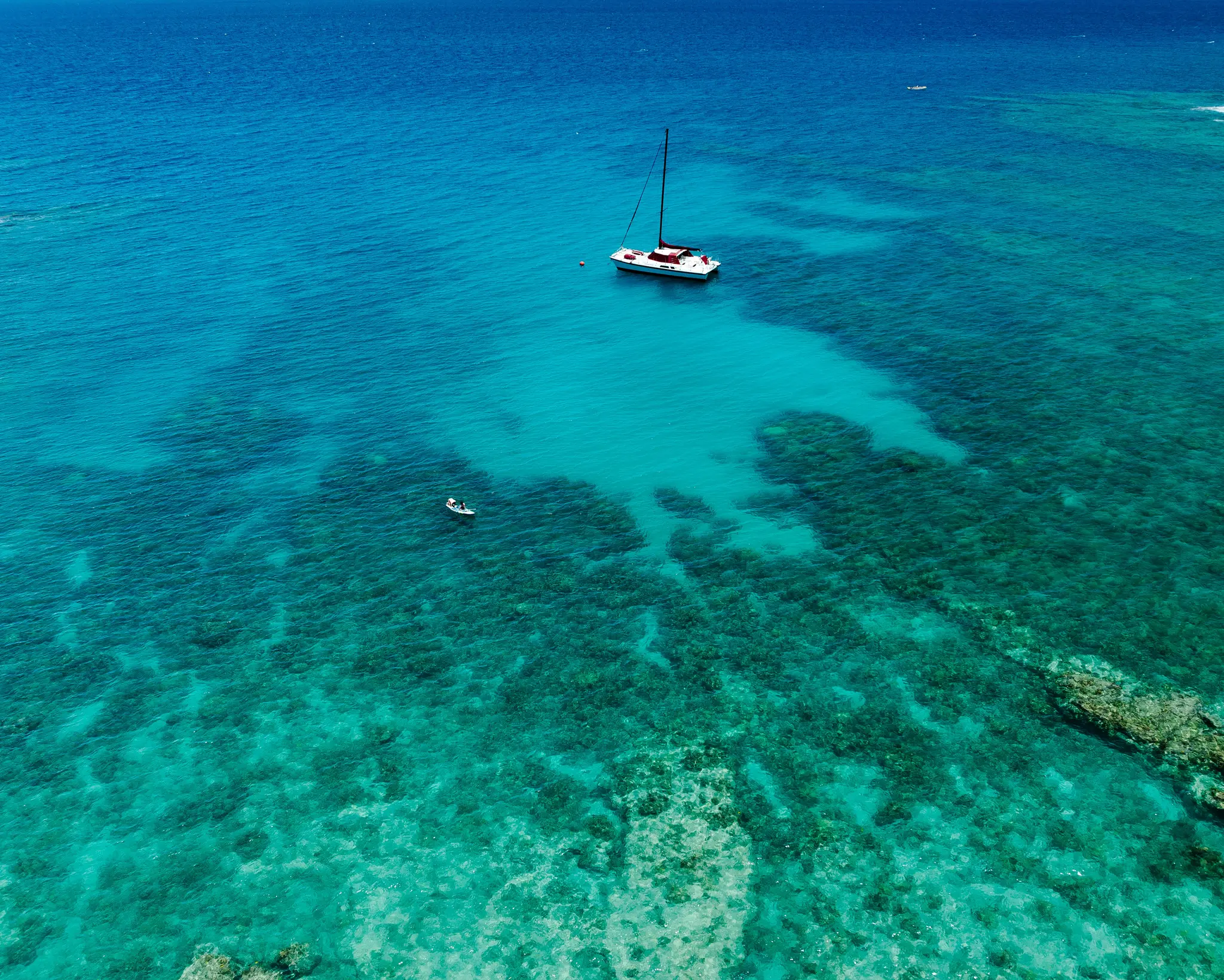 An aerial photo of a white catamaran and a small boat floating on clear turquoise ocean water, with coral reefs visible…