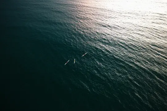 Eine Luftaufnahme zeigt drei kleine Kajaks auf einer weiten Fläche dunkelblauen Wassers, mit einer hellen Lichtreflexion auf