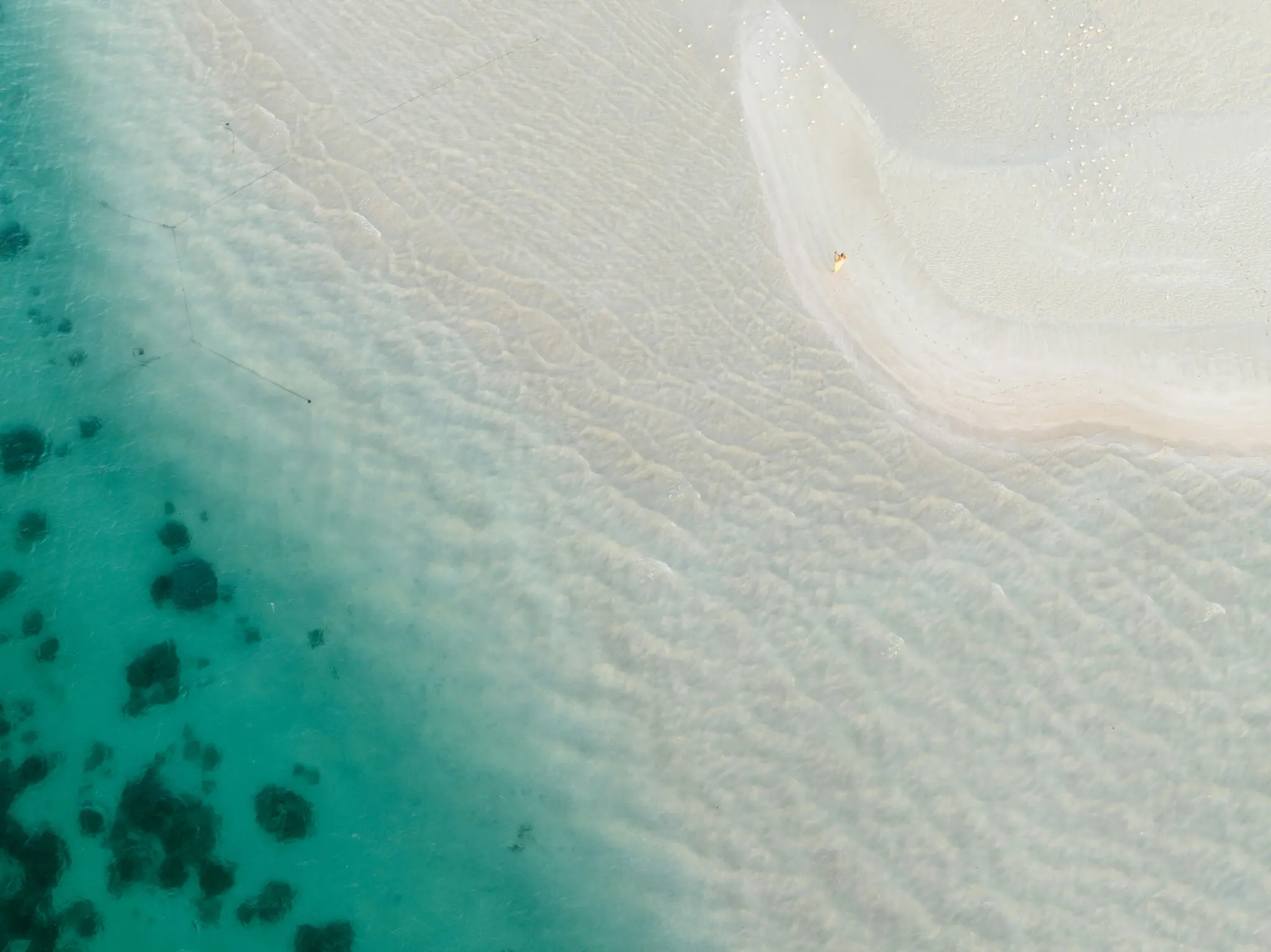 An aerial shot shows a pristine tropical beach. Clear turquoise water with visible ripples meets a curved strip of white…