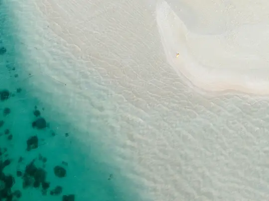An aerial shot shows a pristine tropical beach. Clear turquoise water with visible ripples meets a curved strip of white…