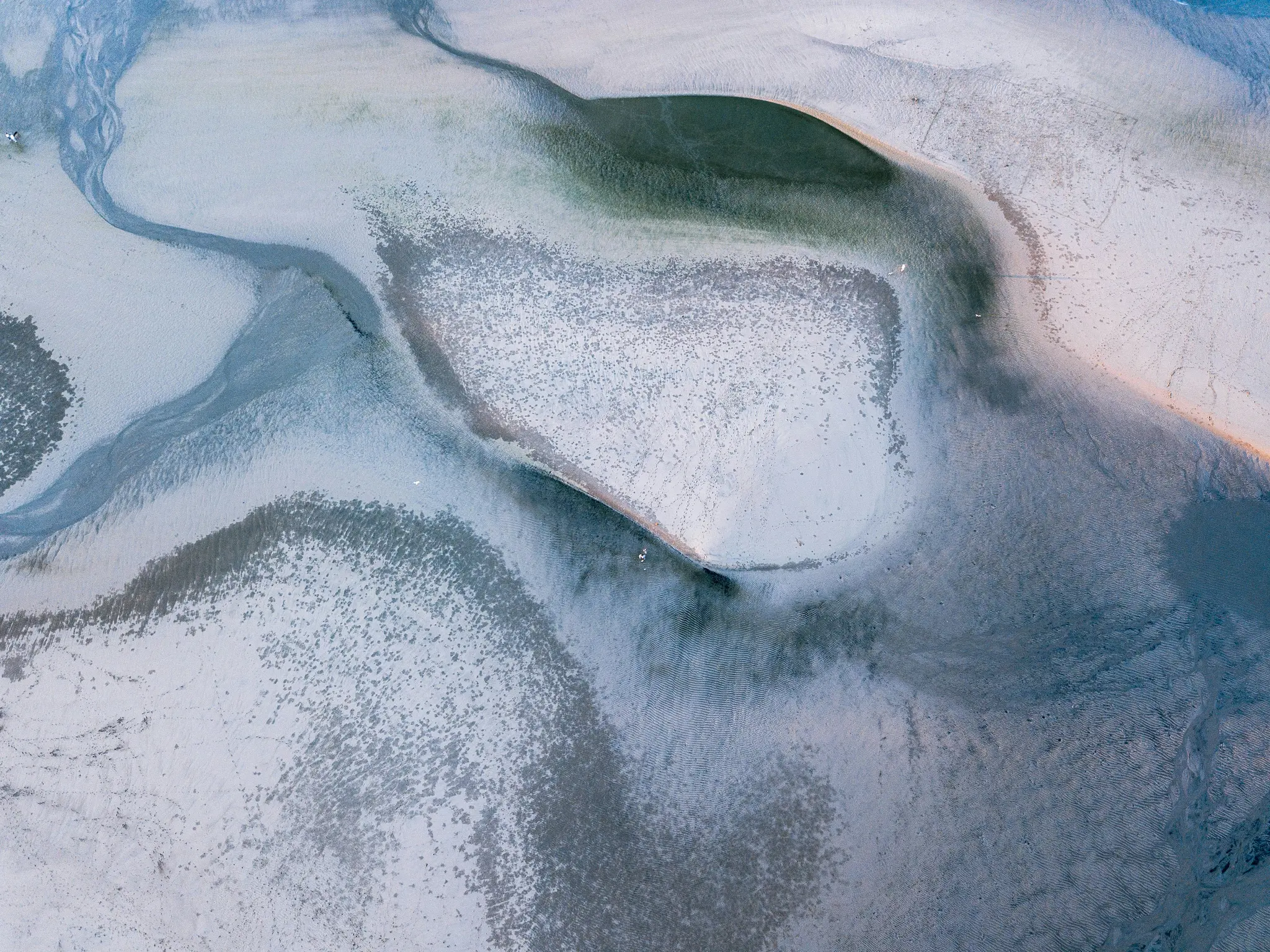 Eine Luftaufnahme einer Wattfläche zeigt komplexe Muster aus hellgrauem Sand und dunkleren blau-grauen Wasserkanälen.