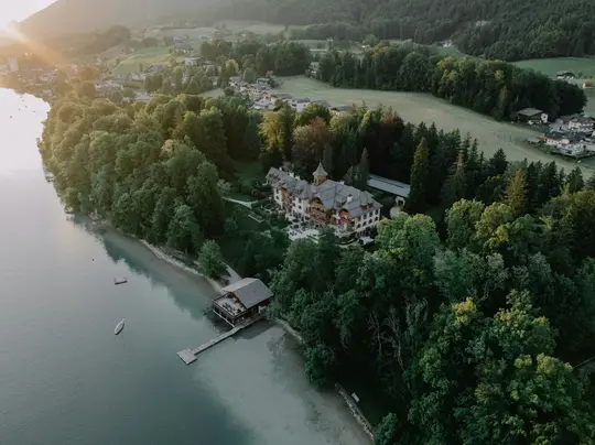 An aerial photo of a grand, multi-story building with intricate architecture, surrounded by lush green trees next to a calm…
