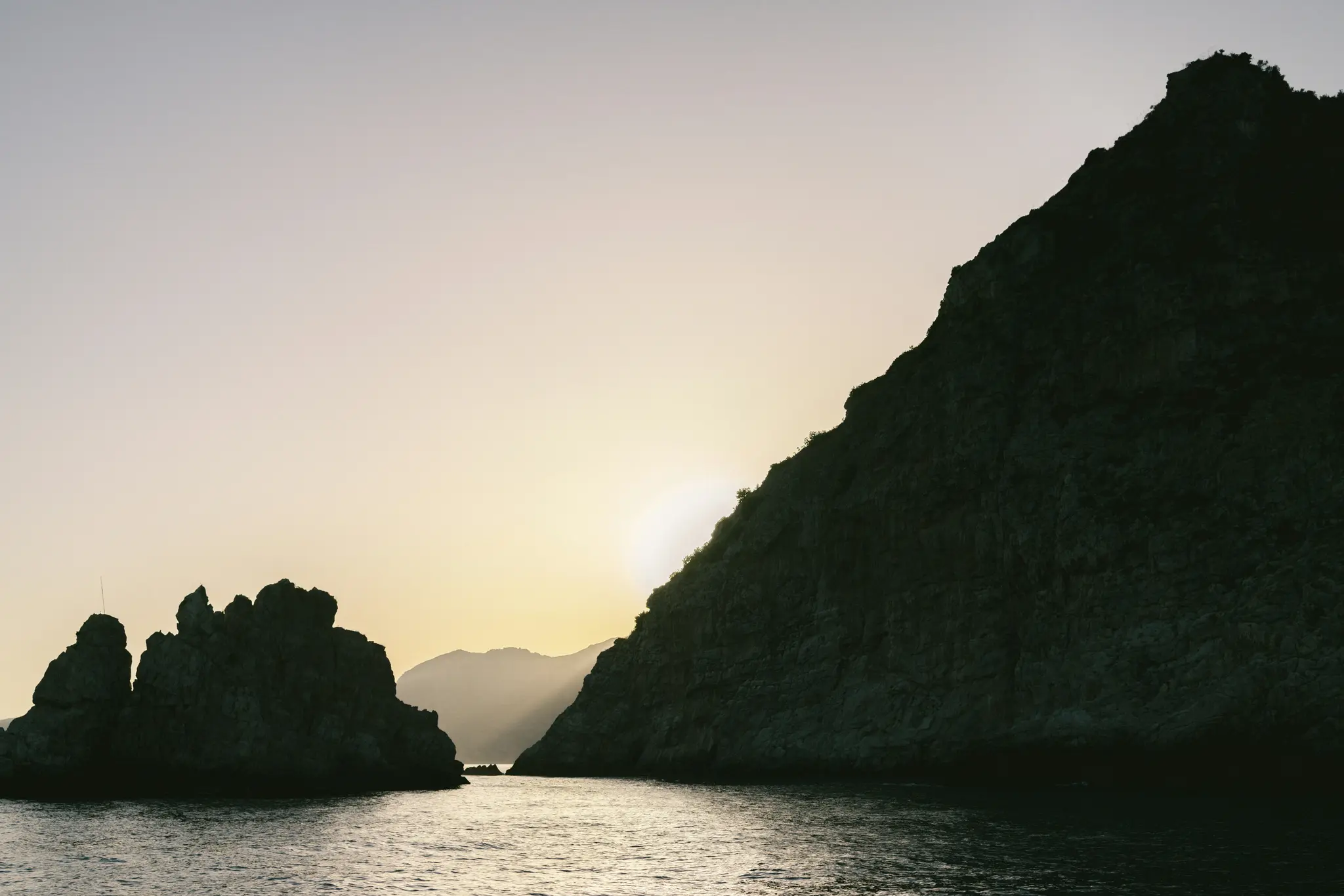 A dramatic landscape photo of dark, jagged rock formations silhouetted against a bright, pale yellow and gray sky at…