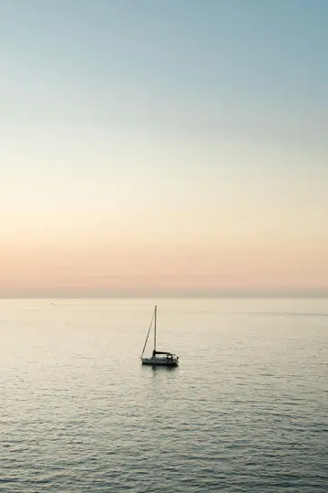 Una foto minimalista di una singola barca a vela con l'albero alzato, ancorata in acque calme grigio-blu sotto un cielo